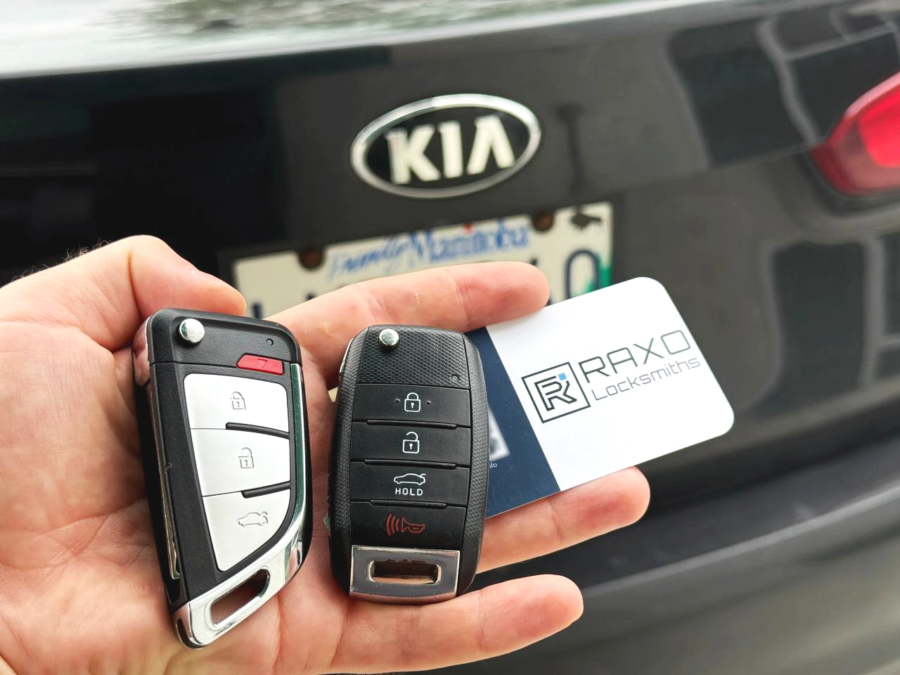 Two replacement Kia flip keys, freshly cut and programmed at the customer's Kia in Winnipeg — RAXO business card on the trunk lid